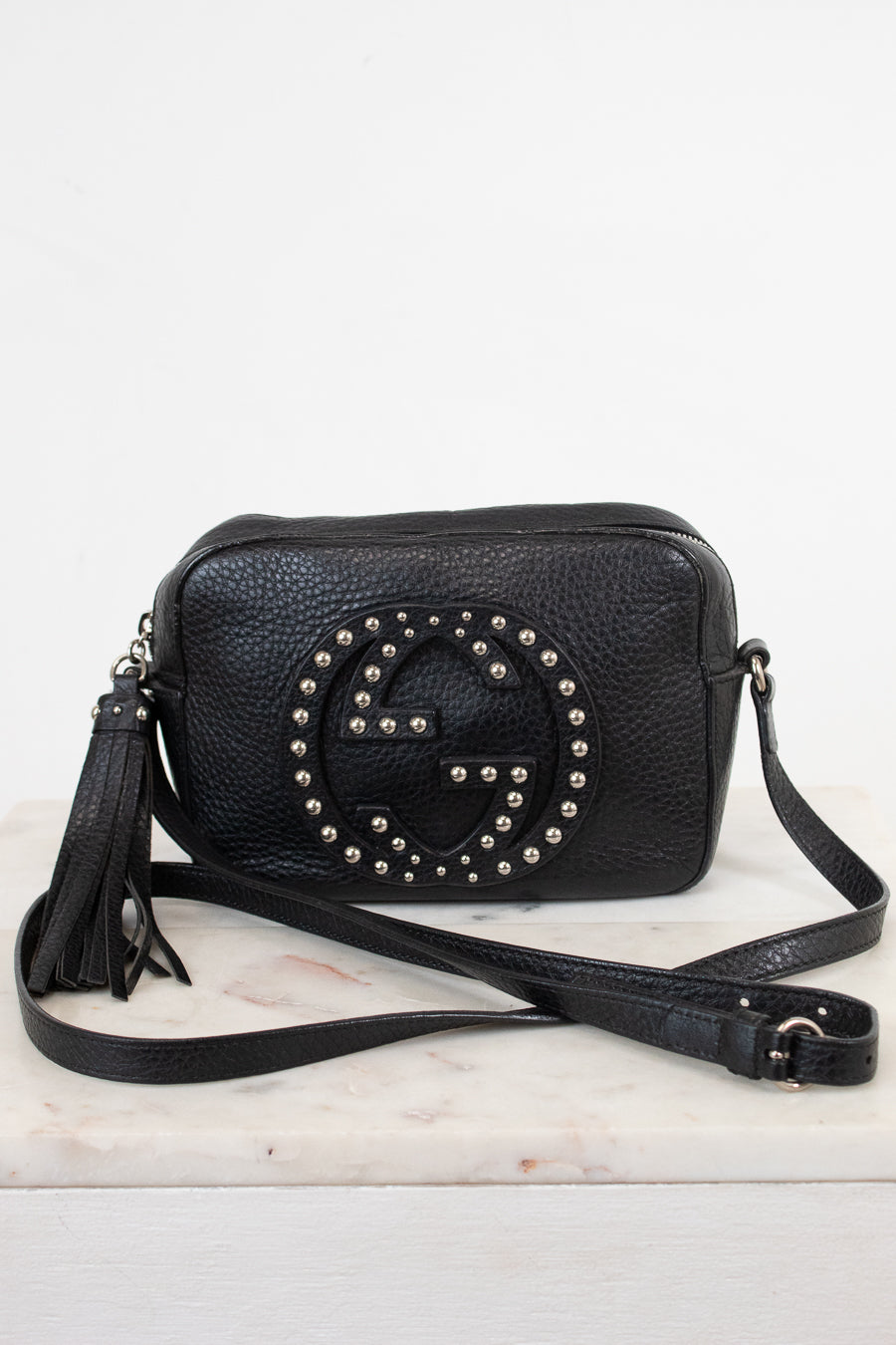A black pebbled leather crossbody bag with a large studded interlocking G logo on the front, resting on a white marble surface against a white background.