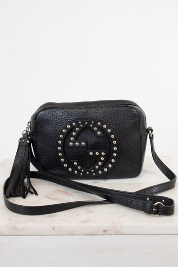 A black pebbled leather crossbody bag with a large studded interlocking G logo on the front, resting on a white marble surface against a white background.