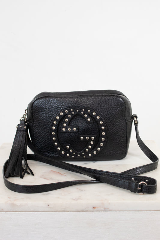 A black pebbled leather crossbody bag with a large studded interlocking G logo on the front, resting on a white marble surface against a white background.