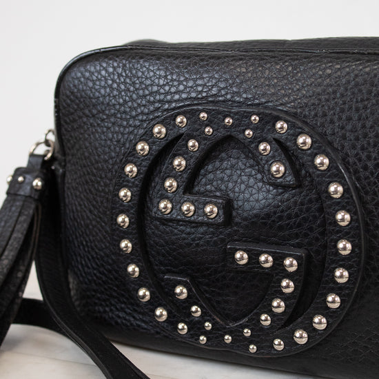 A black pebbled leather crossbody bag featuring a large, circular, studded interlocking G logo on the front, resting on a white marble surface against a white background.