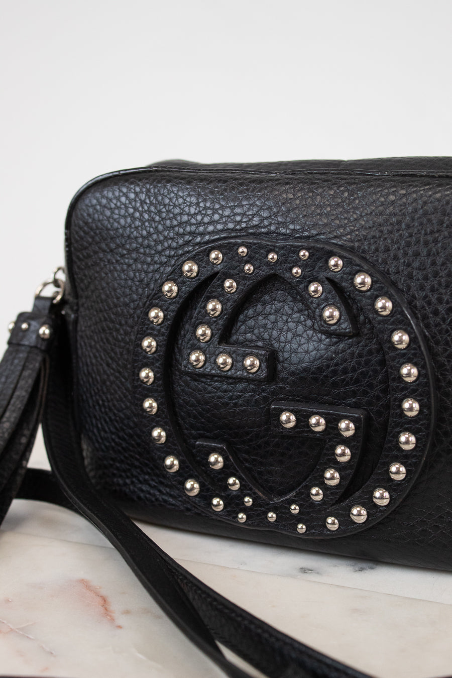 A black pebbled leather crossbody bag featuring a large, circular, studded interlocking G logo on the front, resting on a white marble surface against a white background.