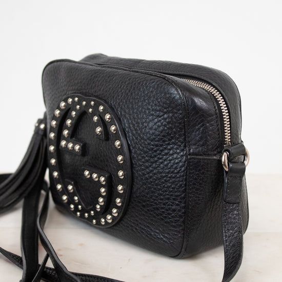 A black pebbled leather crossbody bag with a large circular Gucci logo stud embellishment on the front, resting on a light marble surface against a white background.