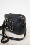 A black pebbled leather crossbody bag with a large circular Gucci logo stud embellishment on the front, resting on a light marble surface against a white background.