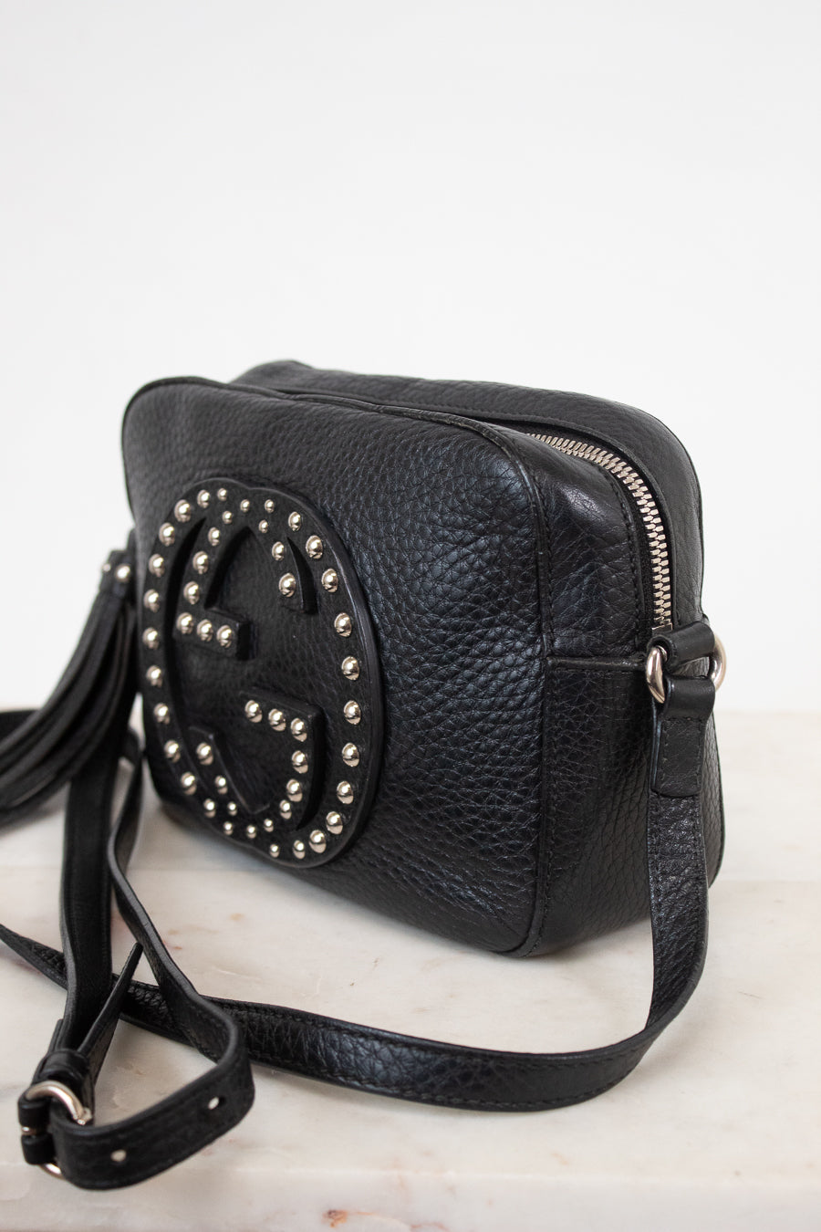 A black pebbled leather crossbody bag with a large circular Gucci logo stud embellishment on the front, resting on a light marble surface against a white background.
