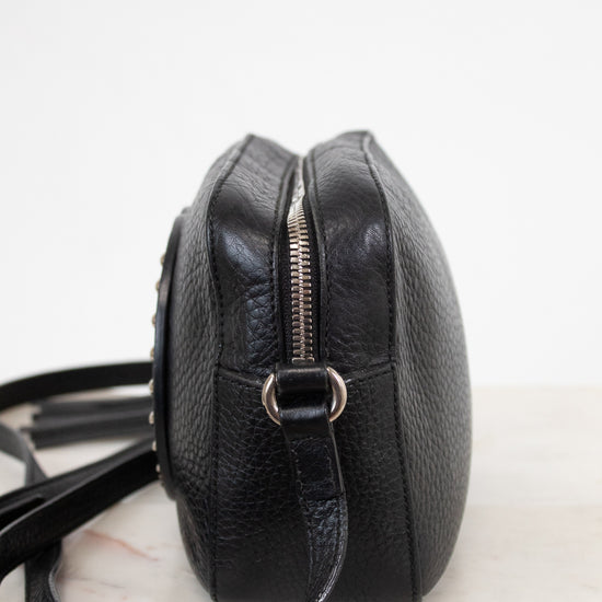 A black pebbled leather crossbody bag with a silver zipper and metal ring, shown from a side profile view on a light marble surface against a white background.