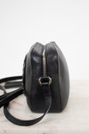 A black pebbled leather crossbody bag with a silver zipper and metal ring, shown from a side profile view on a light marble surface against a white background.