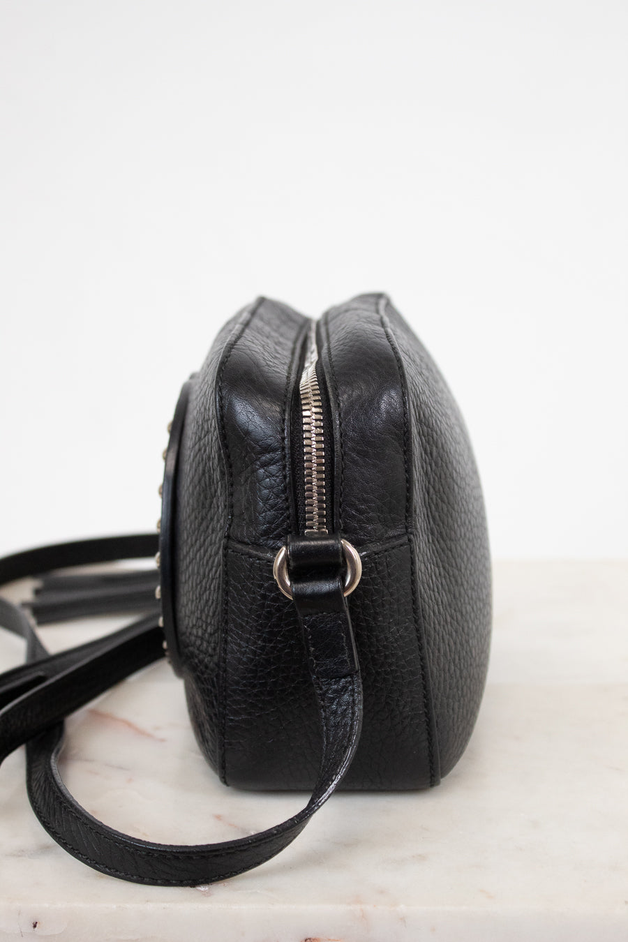 A black pebbled leather crossbody bag with a silver zipper and metal ring, shown from a side profile view on a light marble surface against a white background.