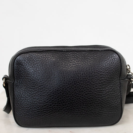 A black pebbled leather crossbody bag with a top zipper, resting on a light marble surface against a white background.