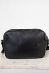 A black pebbled leather crossbody bag with a top zipper, resting on a light marble surface against a white background.