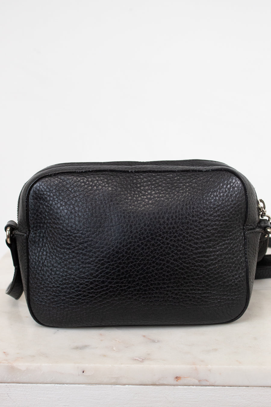 A black pebbled leather crossbody bag with a top zipper, resting on a light marble surface against a white background.