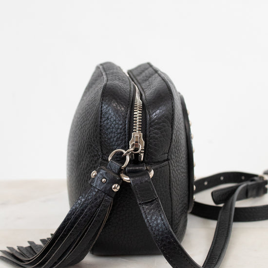 A black pebbled leather crossbody bag with a silver zipper and tassel detail, resting on a light marble surface against a white background.