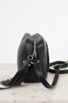 A black pebbled leather crossbody bag with a silver zipper and tassel detail, resting on a light marble surface against a white background.