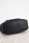 A close-up of a black pebbled leather clutch or small handbag resting on a light marble surface against a white background.