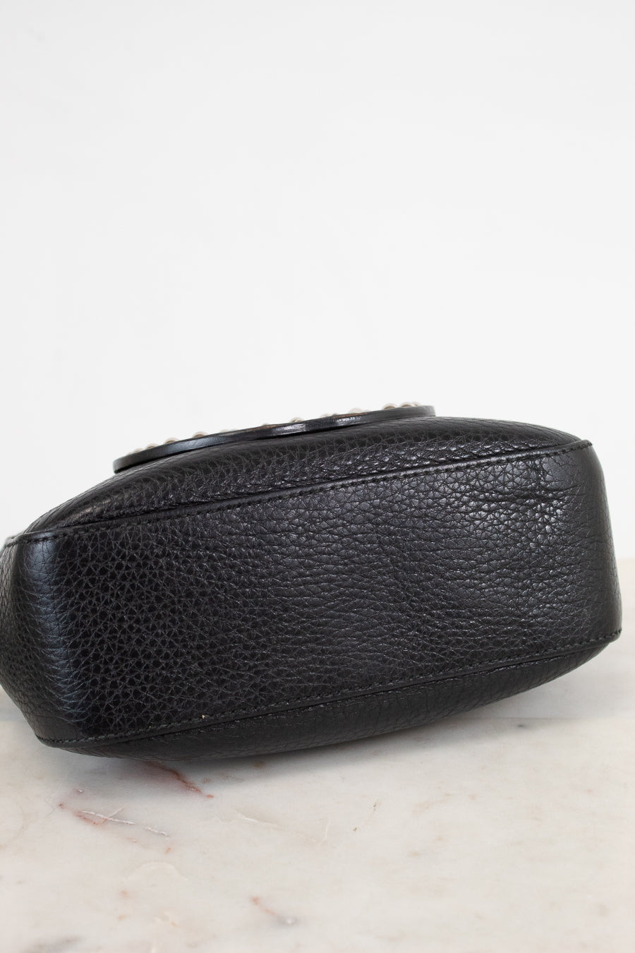 A close-up of a black pebbled leather clutch or small handbag resting on a light marble surface against a white background.
