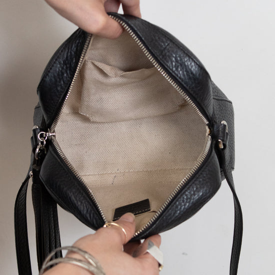 A black textured leather shoulder bag with a beige canvas interior, shown open with a person's hands holding it against a plain light background.