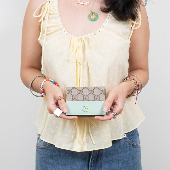A woman wearing a pale yellow ruffled sleeveless top and blue jeans holds a Gucci monogram wallet in mint green and pale yellow. She is wearing several gold necklaces and bracelets against a plain light gray background.