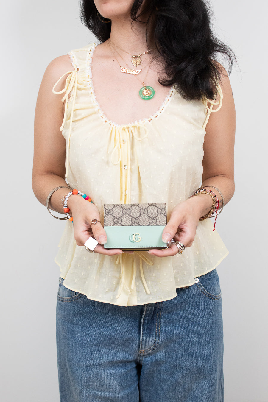 A woman wearing a pale yellow ruffled sleeveless top and blue jeans holds a Gucci monogram wallet in mint green and pale yellow. She is wearing several gold necklaces and bracelets against a plain light gray background.