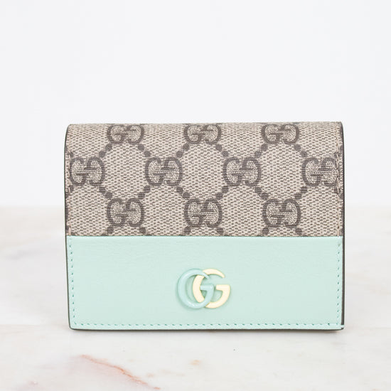 A Gucci wallet in beige GG Supreme canvas with a light mint green leather lower section and a gold-tone interlocking G logo on the front, set against a white background.