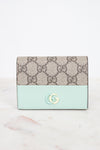 A Gucci wallet in beige GG Supreme canvas with a light mint green leather lower section and a gold-tone interlocking G logo on the front, set against a white background.
