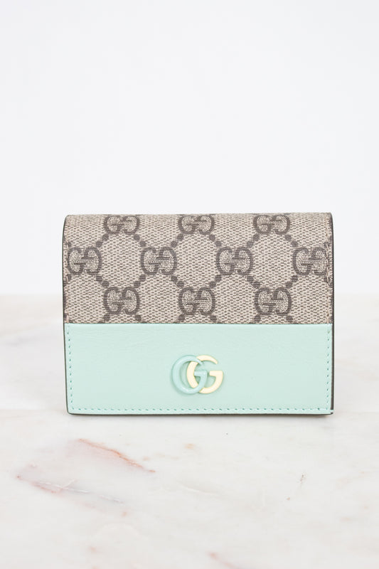A Gucci wallet in beige GG Supreme canvas with a light mint green leather lower section and a gold-tone interlocking G logo on the front, set against a white background.