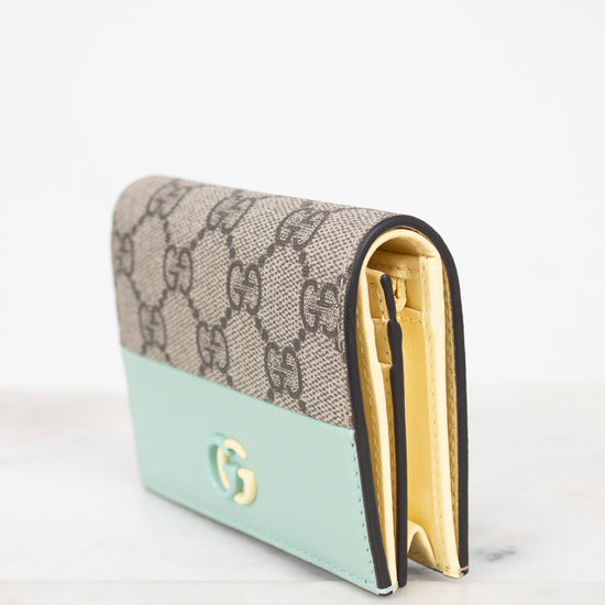 A Gucci wallet in the brand's signature GG Supreme canvas with a mint green leather lower section and gold-tone hardware, displayed on a light marble surface against a white background.
