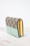 A Gucci wallet in the brand's signature GG Supreme canvas with a mint green leather lower section and gold-tone hardware, displayed on a light marble surface against a white background.