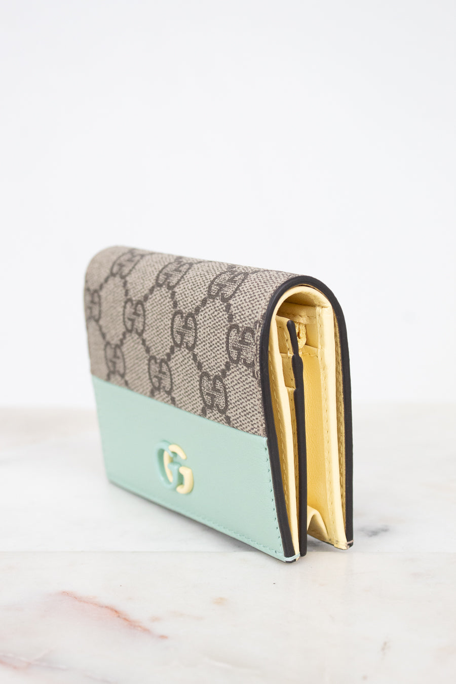 A Gucci wallet in the brand's signature GG Supreme canvas with a mint green leather lower section and gold-tone hardware, displayed on a light marble surface against a white background.