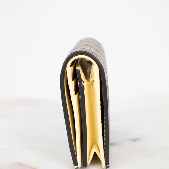 A close-up side view of a slim bifold wallet standing upright on a light marble surface against a white background. The wallet has a textured beige monogram exterior with a bright yellow interior lining.