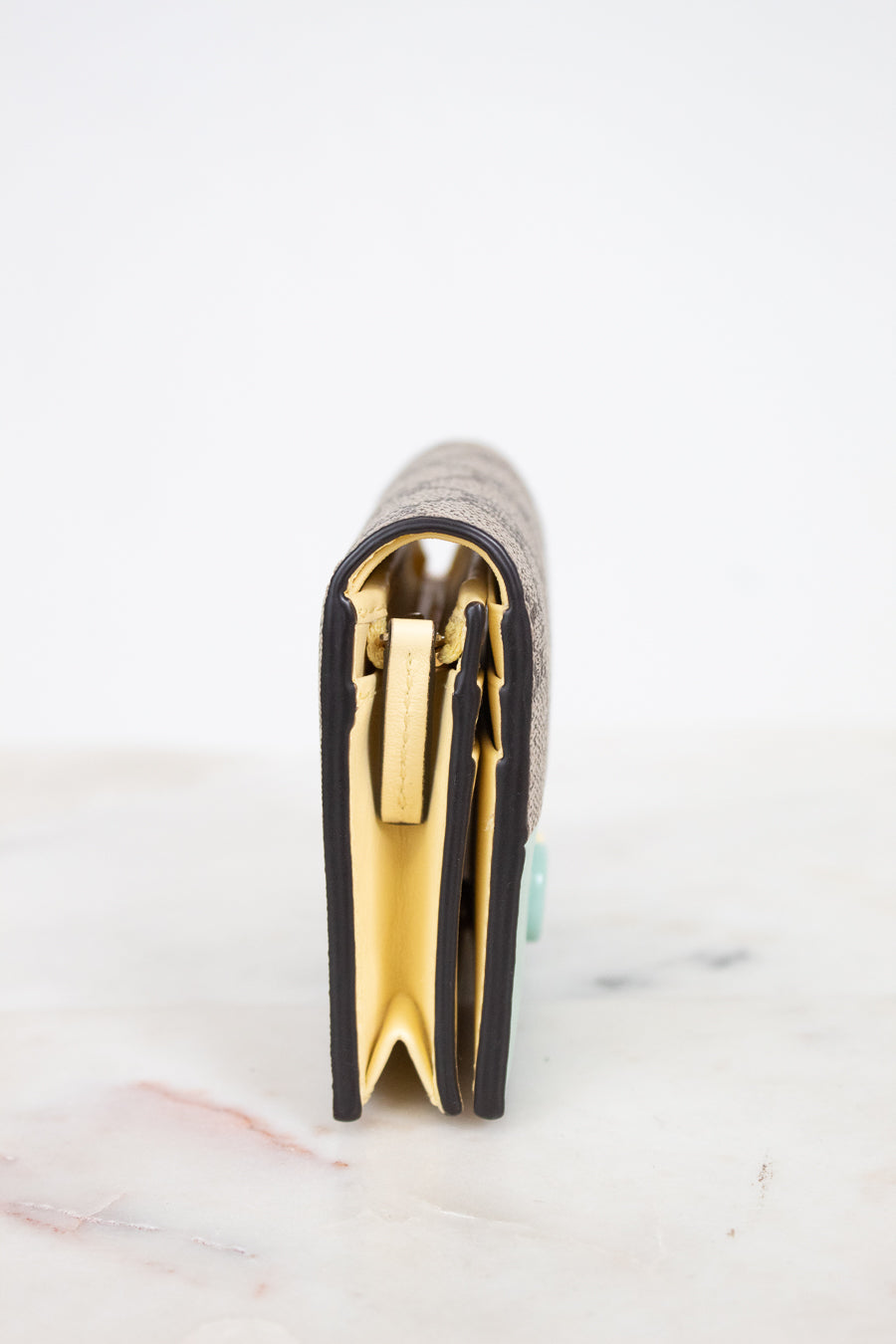 A close-up side view of a wallet standing upright on a light marble surface against a white background, showing a textured exterior, black piping trim, and prominent pale yellow interior.