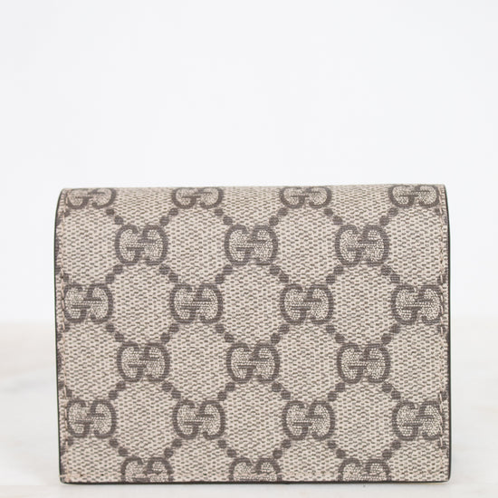 View of back of small Gucci wallet in beige GG Supreme canvas with a dark and beige monogram pattern, resting on a white marble surface against a white background.