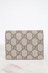 View of back of small Gucci wallet in beige GG Supreme canvas with a dark and beige monogram pattern, resting on a white marble surface against a white background.