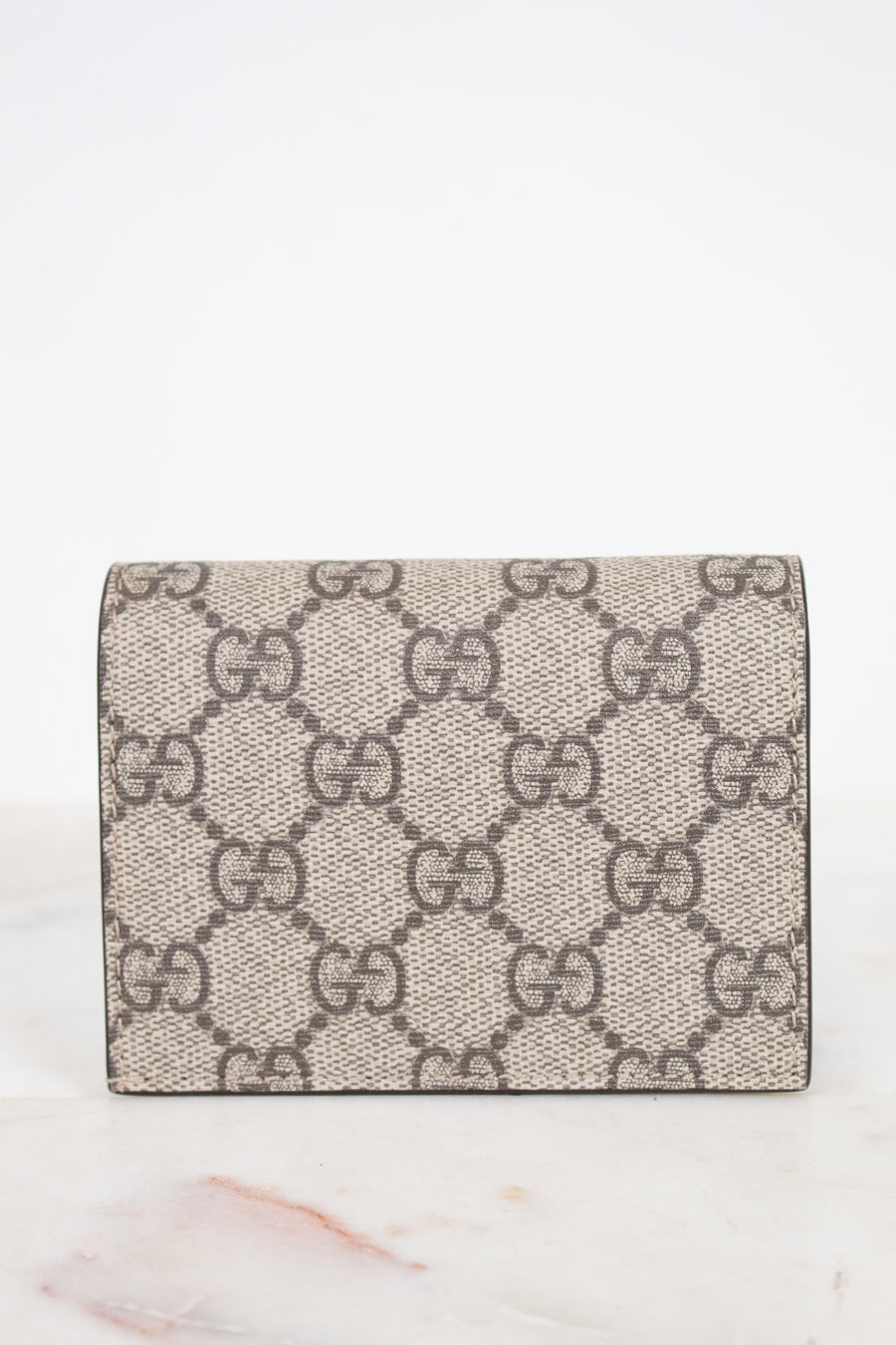 View of back of small Gucci wallet in beige GG Supreme canvas with a dark and beige monogram pattern, resting on a white marble surface against a white background.