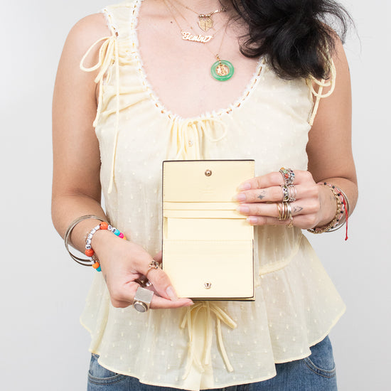 A woman wearing a pale yellow ruffled sleeveless top and blue jeans holds a small pale yellow-colored leather wallet or clutch with a gold-tone snap button and card slots.
