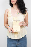 A woman wearing a pale yellow ruffled sleeveless top and blue jeans holds a small pale yellow-colored leather wallet or clutch with a gold-tone snap button and card slots.