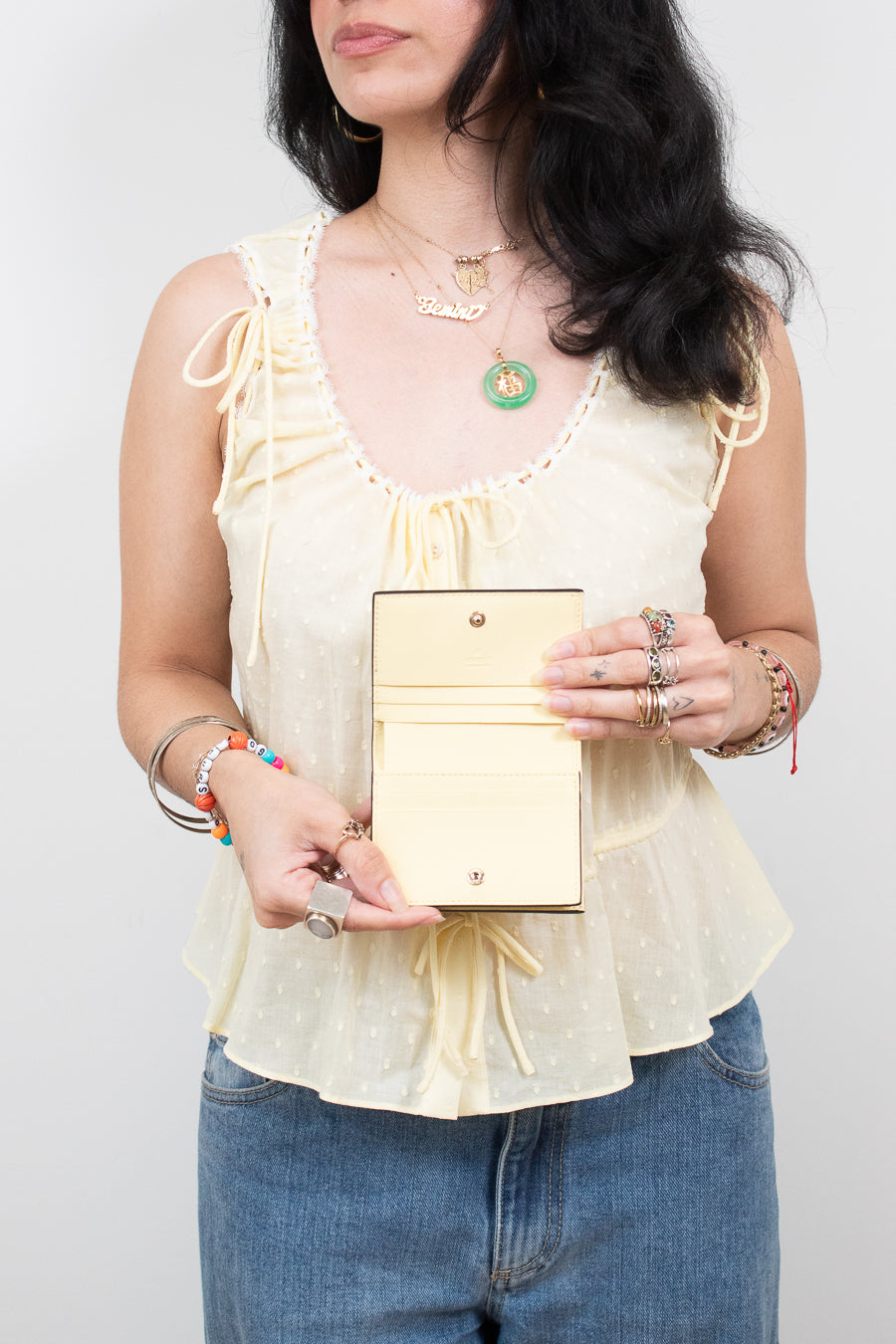 A woman wearing a pale yellow ruffled sleeveless top and blue jeans holds a small pale yellow-colored leather wallet or clutch with a gold-tone snap button and card slots.