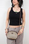 A woman wearing a black ribbed tank top and gray sweatpants holds a small Gucci monogram crossbody bag with a brown strap against a plain light gray background.