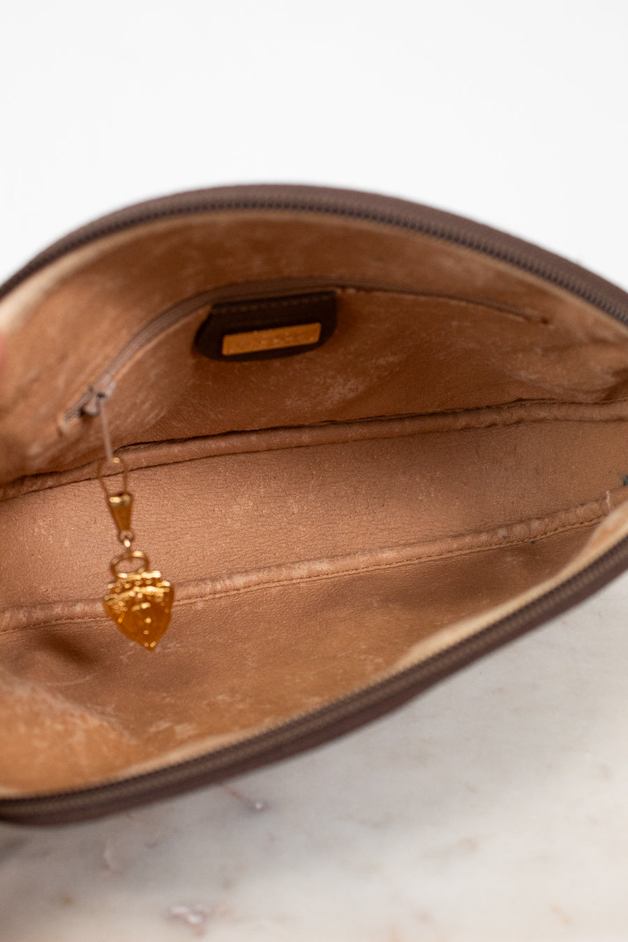Close-up of the interior of a brown zippered pouch or clutch, showing a tan satin lining, a small interior label, and a dangling gold zipper pull.