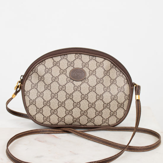 A vintage Gucci oval shoulder bag in the classic GG Supreme canvas, featuring a brown leather trim and strap, resting on a white marble surface against a white background.