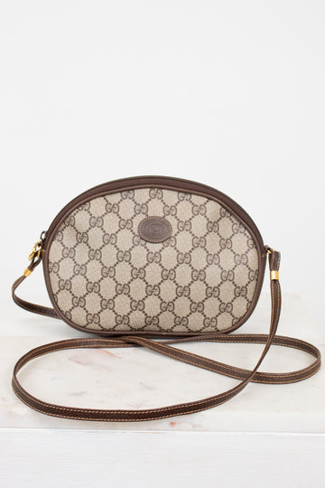 A vintage Gucci oval shoulder bag in the classic GG Supreme canvas, featuring a brown leather trim and strap, resting on a white marble surface against a white background.