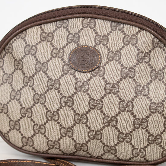 A close-up of a Gucci monogram canvas shoulder bag in beige and brown, featuring the classic GG pattern, a brown leather trim, and a central round Gucci logo patch. A matching brown strap is visible in the foreground against a light background.
