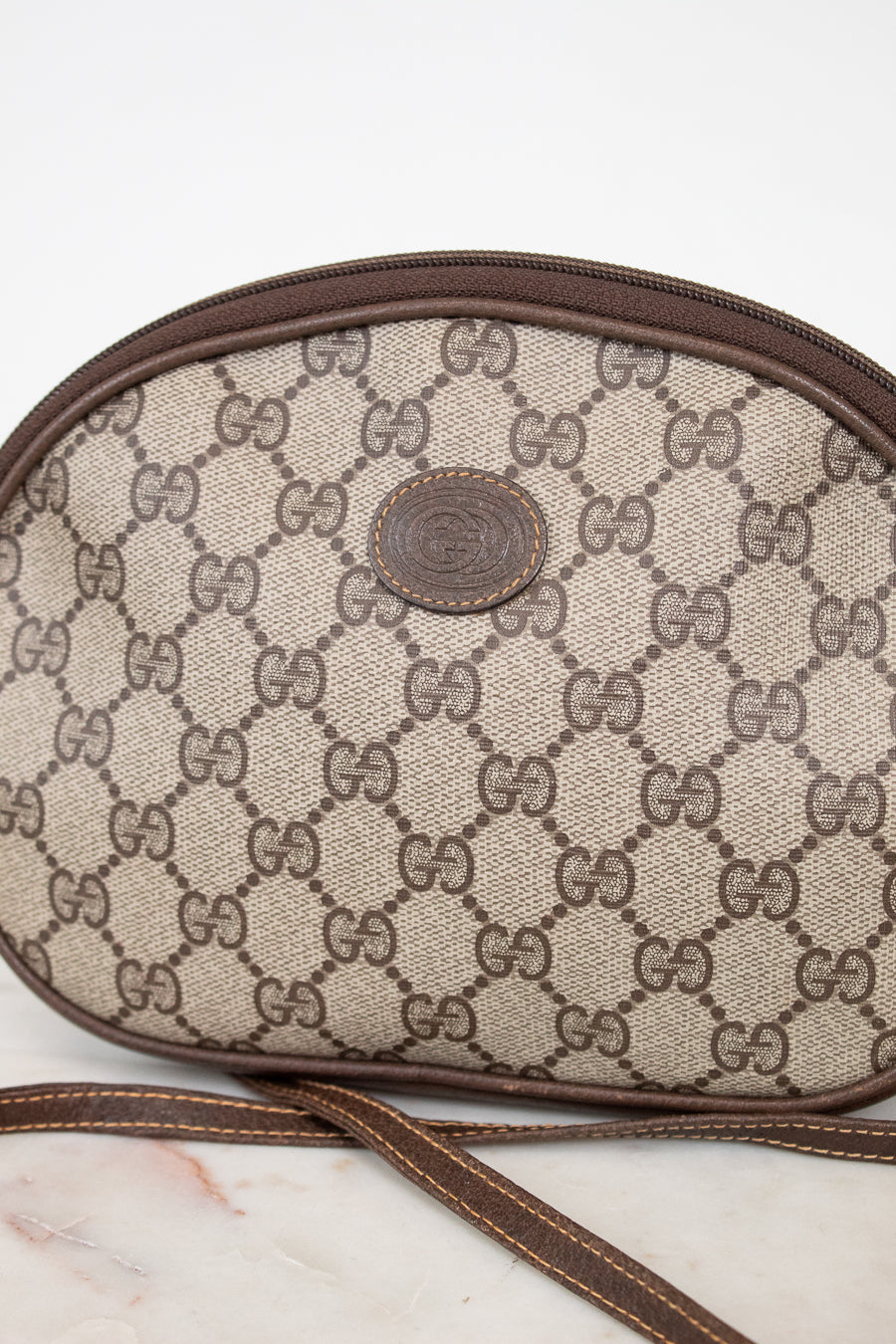 A close-up of a Gucci monogram canvas shoulder bag in beige and brown, featuring the classic GG pattern, a brown leather trim, and a central round Gucci logo patch. A matching brown strap is visible in the foreground against a light background.