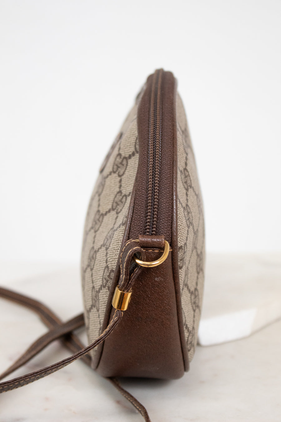 A vintage Gucci monogram canvas pouch with brown leather trim and gold-tone hardware, shown from a side profile view on a light marble surface.