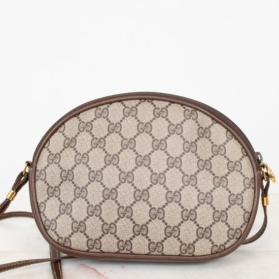 A Gucci GG Supreme oval shoulder bag in beige and brown monogram canvas with brown leather trim and gold-tone hardware, resting on a light marble surface against a white background.