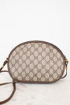 A Gucci GG Supreme oval shoulder bag in beige and brown monogram canvas with brown leather trim and gold-tone hardware, resting on a light marble surface against a white background.