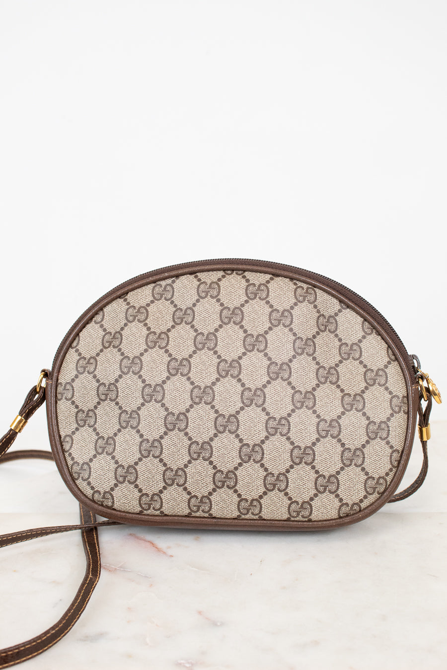 A Gucci GG Supreme oval shoulder bag in beige and brown monogram canvas with brown leather trim and gold-tone hardware, resting on a light marble surface against a white background.