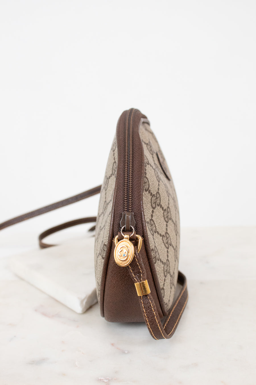 A Gucci shoulder bag in beige GG Supreme canvas with brown leather trim and gold-tone hardware, shown from a side profile on a light marble surface against a white background.