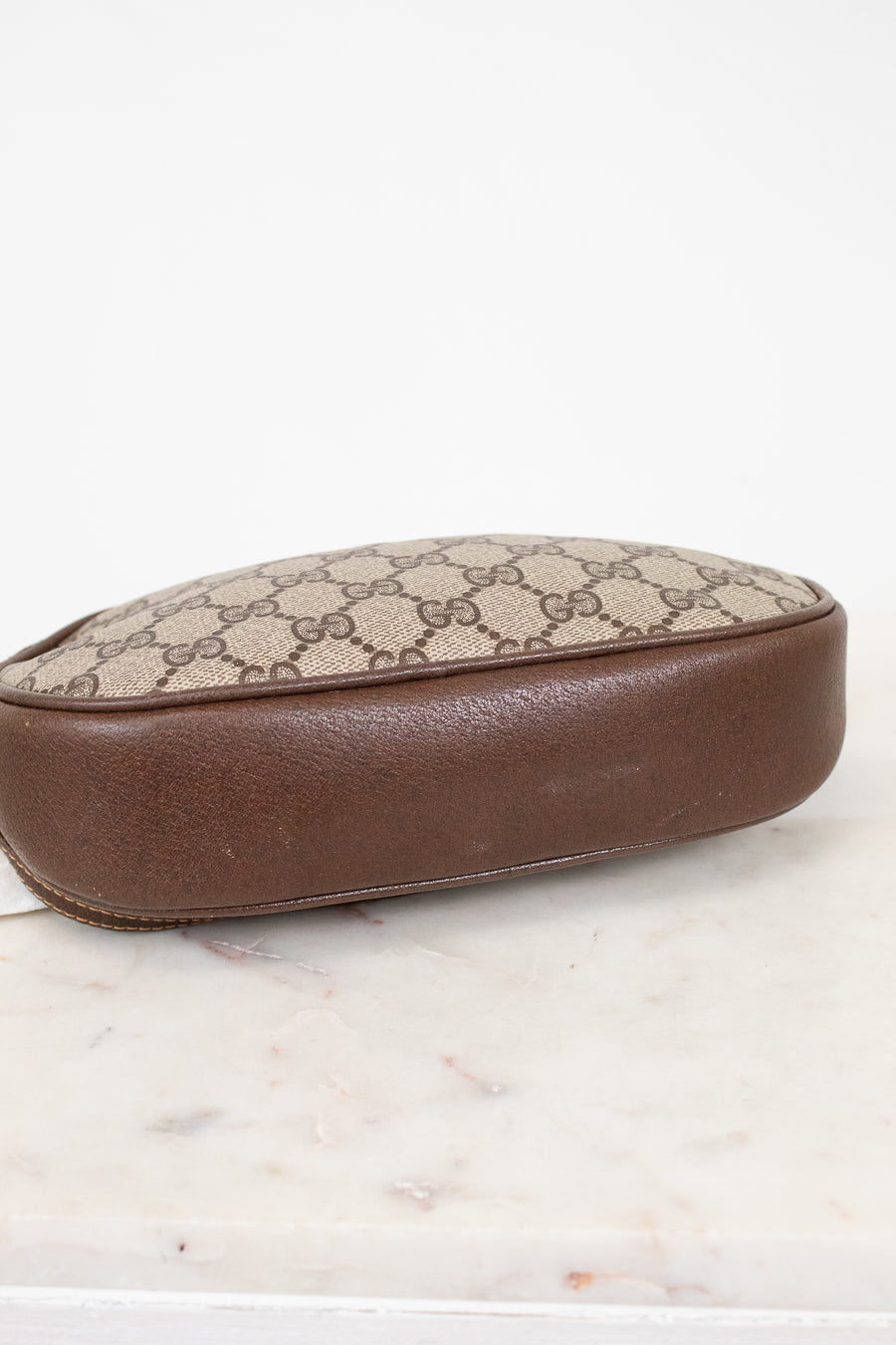 A vintage Gucci pouch featuring the classic GG Supreme canvas in beige and brown, finished with a smooth brown leather base, shown on a white marble surface against a white background.