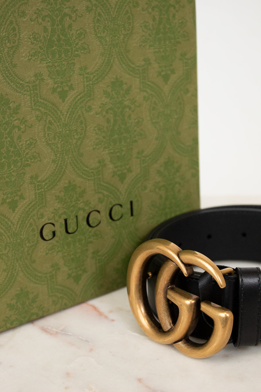Gucci belt with gold buckle on a marble surface next to a Gucci box.