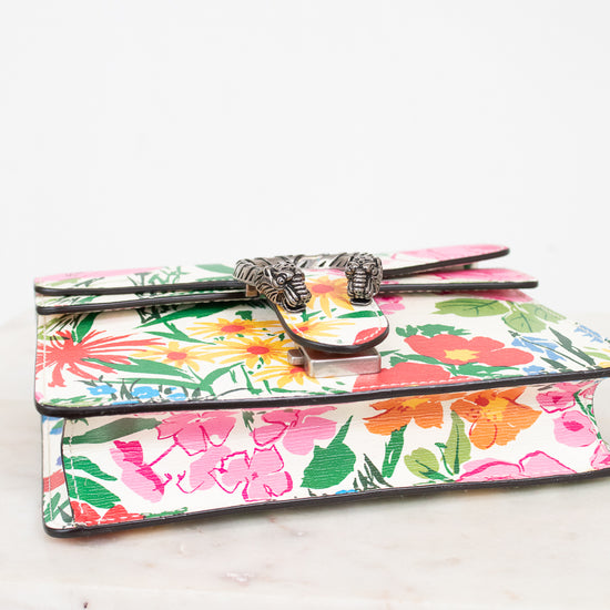 A small leather clutch wallet with a colorful tropical floral print, featuring a silver-toned metal clasp closure, resting on a white marble surface against a white background.