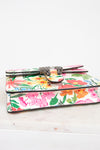A small leather clutch wallet with a colorful tropical floral print, featuring a silver-toned metal clasp closure, resting on a white marble surface against a white background.
